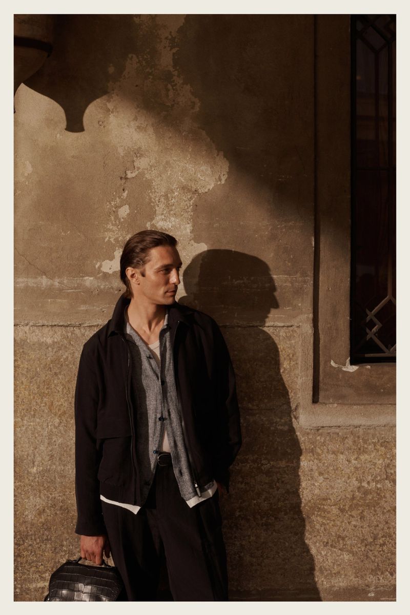 Adult male model wearing a black jacket and holding a bag, posing against an aged textured wall with a window shadow.