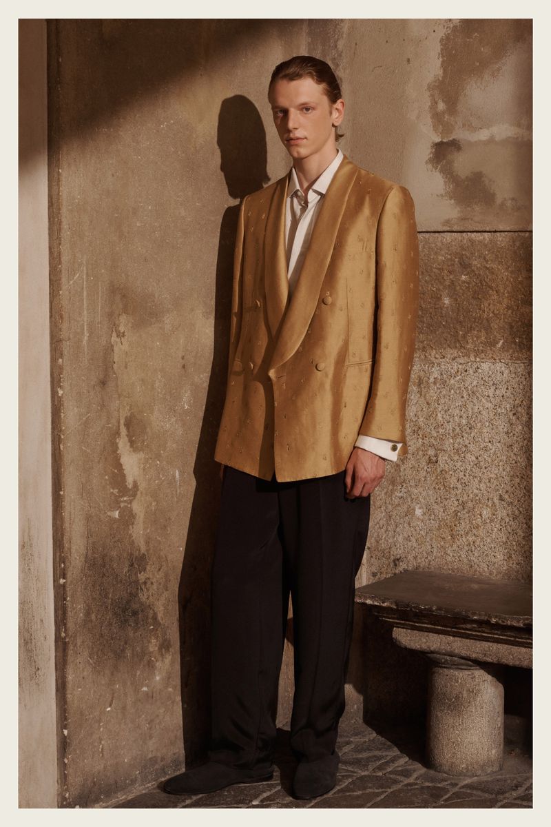 Male fashion model poses in a tan blazer and black pants against a textured wall, lit by natural light creating a shadow.