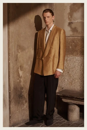 Male fashion model poses in a tan blazer and black pants against a textured wall, lit by natural light creating a shadow.