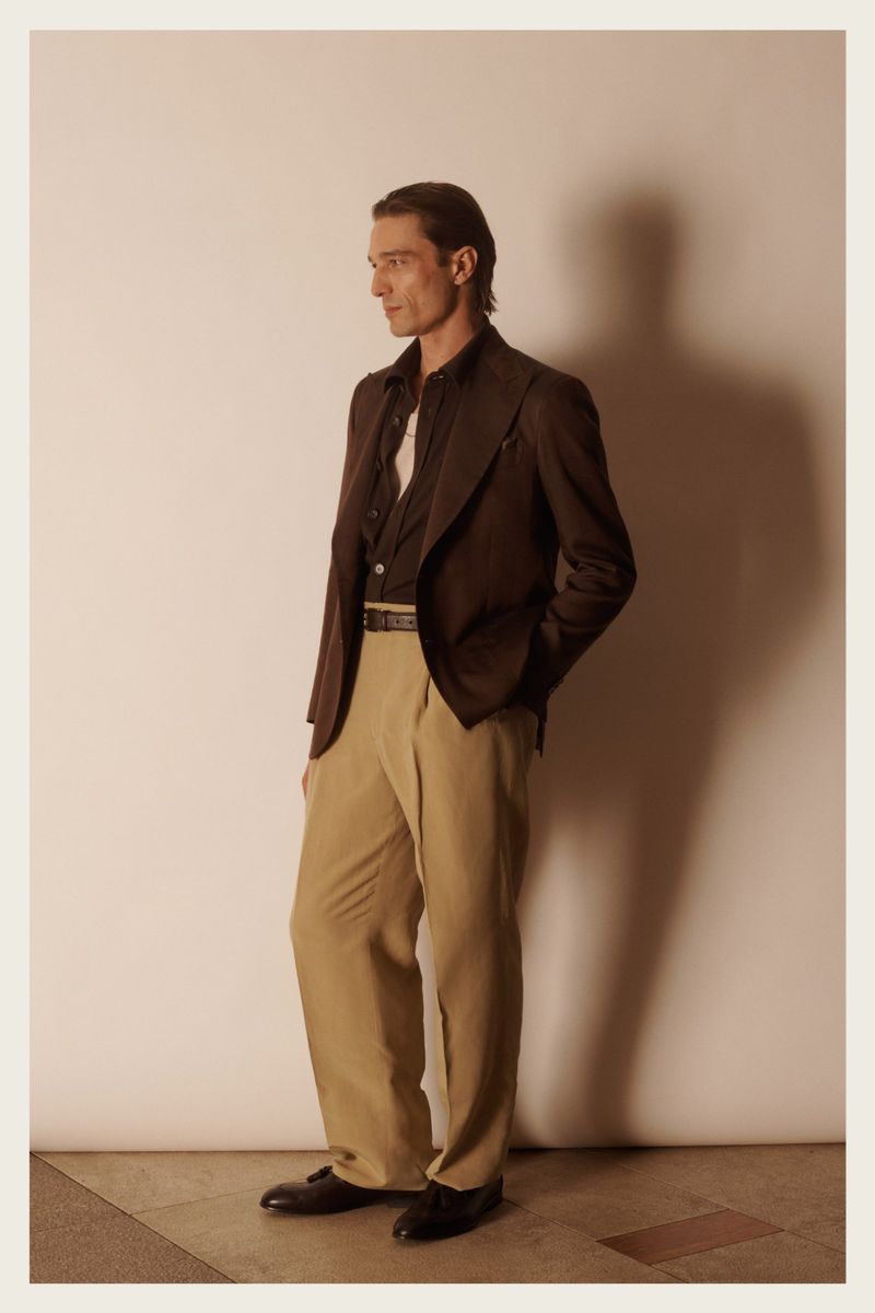 Stylish adult male fashion model poses in a brown blazer, dark shirt, and tan dress pants against a neutral studio backdrop.