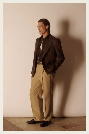 Stylish adult male fashion model poses in a brown blazer, dark shirt, and tan dress pants against a neutral studio backdrop.