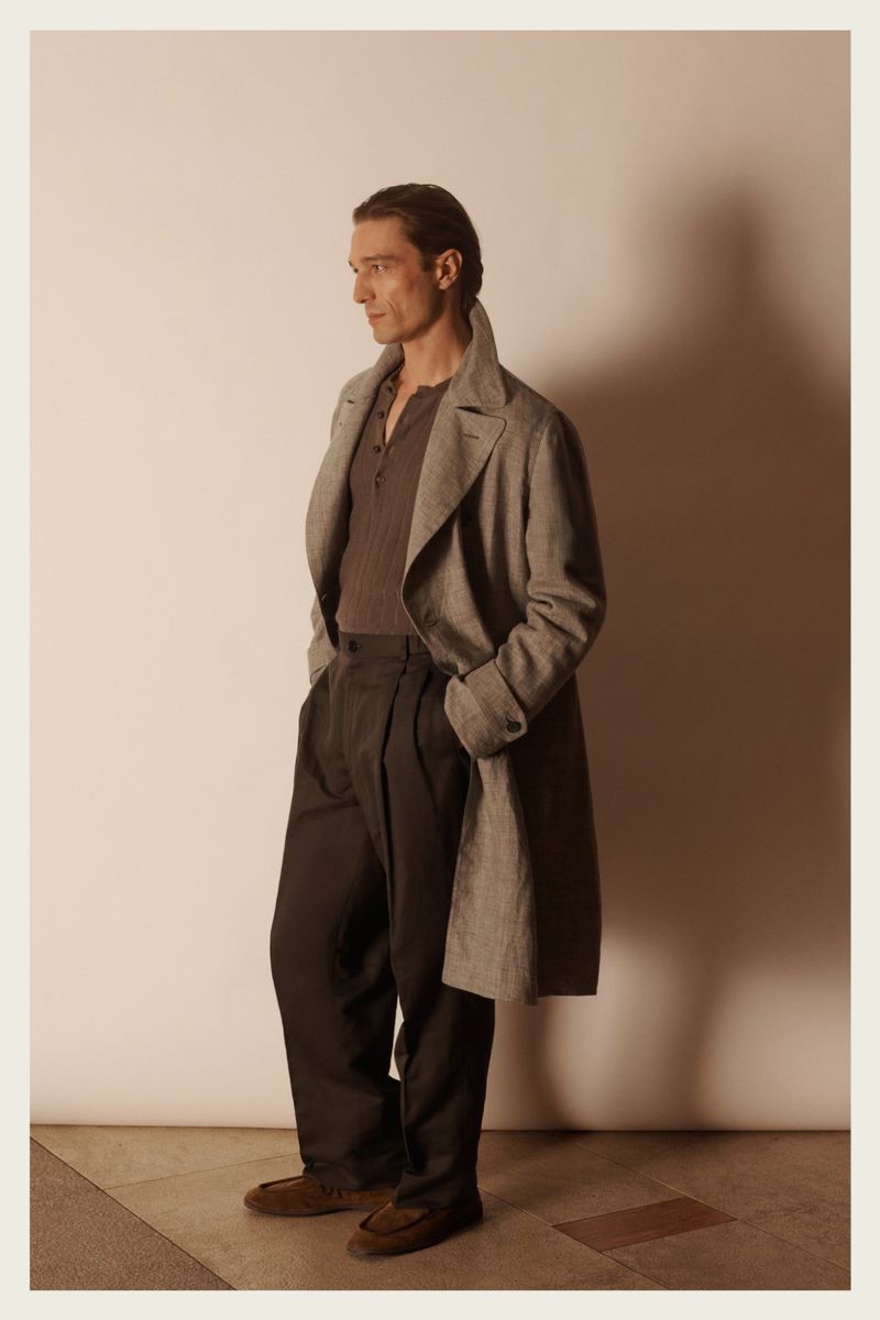 A man in a grey coat and dark pants poses in a studio with soft lighting. He is captured in a medium shot, showcasing fashion attire.