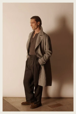 A man in a grey coat and dark pants poses in a studio with soft lighting. He is captured in a medium shot, showcasing fashion attire.
