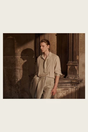 An adult man stands near a carved stone wall, wearing a beige linen shirt and pants, in soft lighting and a medium shot.