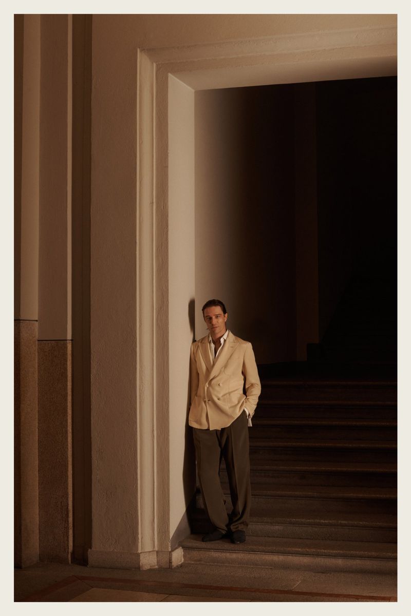 A man in a beige blazer and olive pants stands near a doorway in a brightly lit interior. Classic and fashionable.