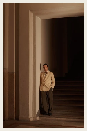 A man in a beige blazer and olive pants stands near a doorway in a brightly lit interior. Classic and fashionable.