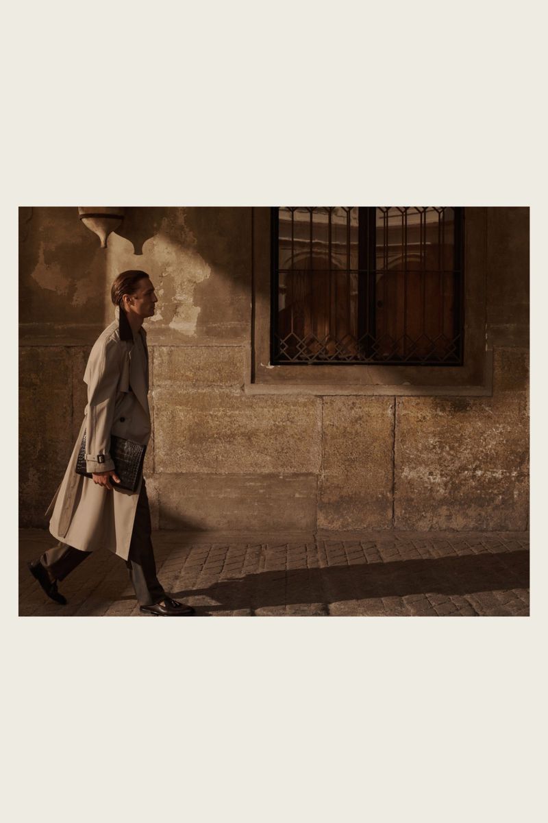 A woman in a trench coat walks past an aged stone building with a barred window in urban setting with warm lighting.