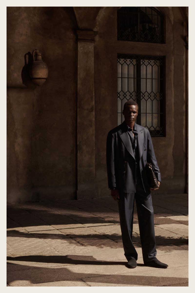An elegant Black male model poses outdoors wearing a blue suit and holding a satchel, set against a weathered building with shadows.