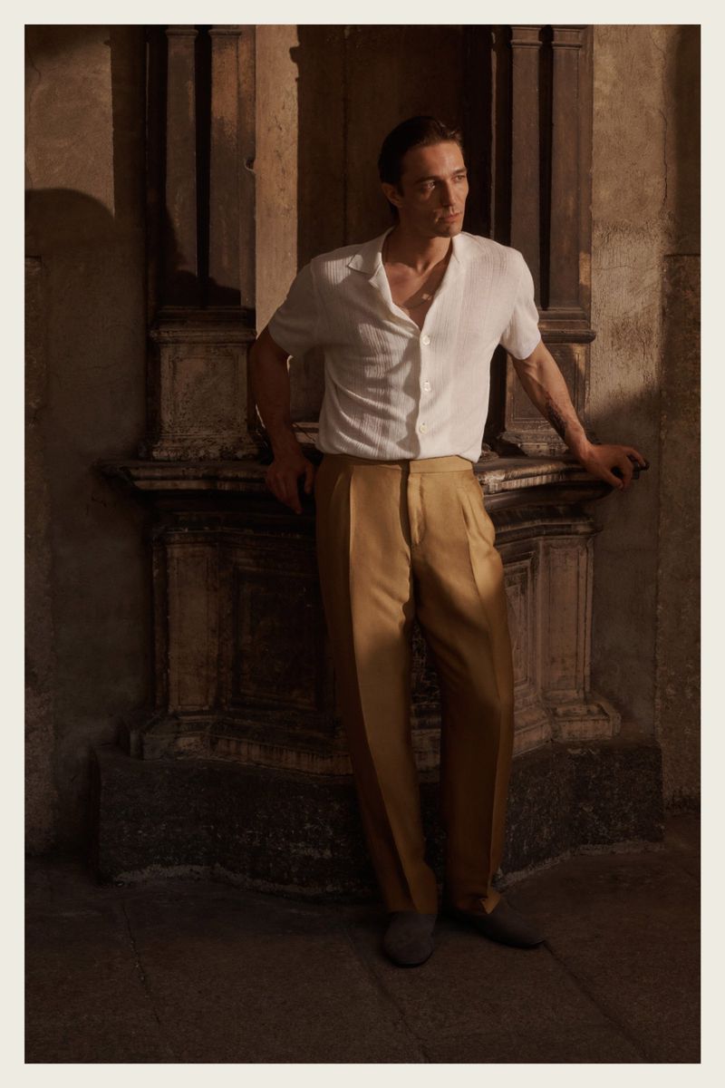 Adult male fashion model poses in a white short-sleeved shirt and tan dress pants while leaning against a stone feature indoors.
