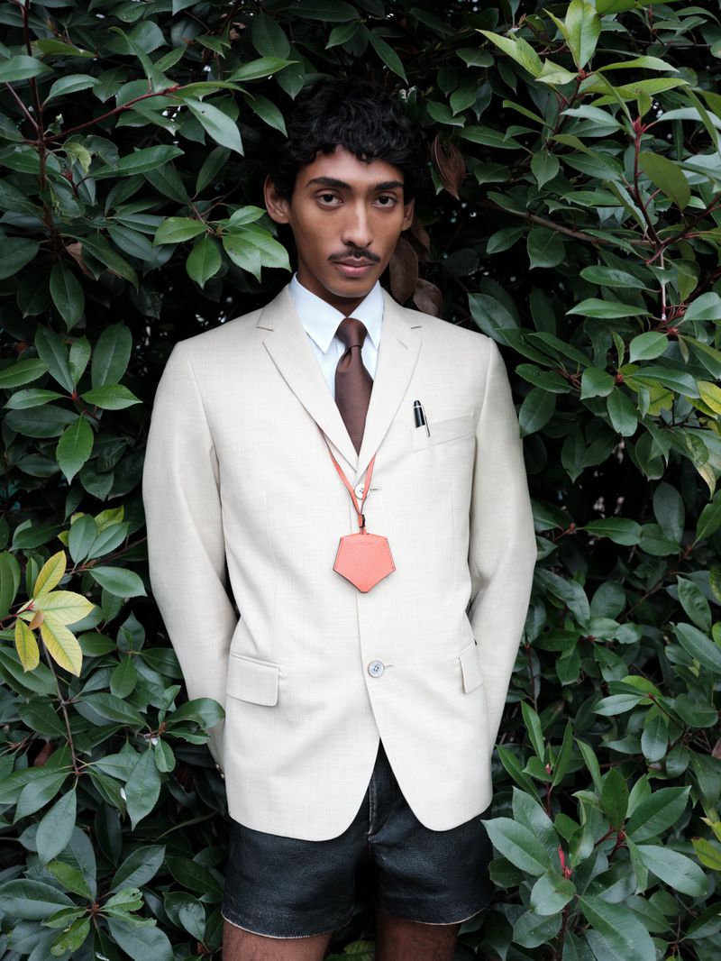 Viknes Waren wears a cream tailored blazer, brown tie, and black leather shorts standing against lush green foliage.