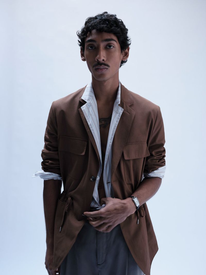 South Asian male model wearing a brown jacket over a striped shirt against a plain white background