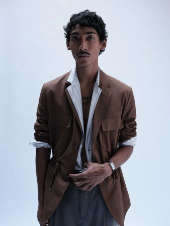 South Asian male model wearing a brown jacket over a striped shirt against a plain white background