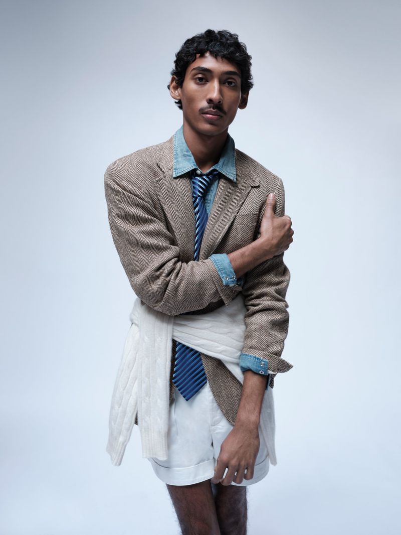 Viknes Waren posing in a tweed blazer, denim shirt, blue striped tie, and white shorts against a pale seamless backdrop.