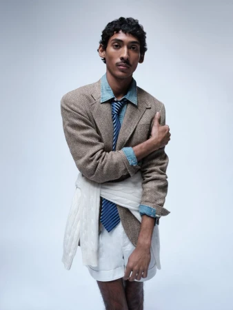 Viknes Waren posing in a tweed blazer, denim shirt, blue striped tie, and white shorts against a pale seamless backdrop.