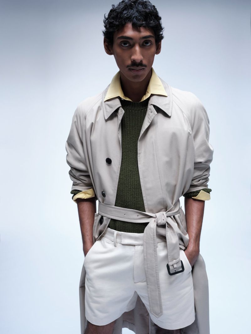 Model Viknes Waren posing in a belted beige trench coat over an olive green knit sweater and white shorts against a light grey studio backdrop.