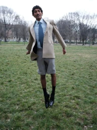 Model Viknes Waren jumping mid-air on a grassy lawn wearing a beige double-breasted blazer, grey shorts, and blue tie.