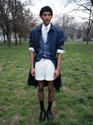 Model Viknes Waren posing on a grassy lawn wearing a navy blue trench coat, layered waistcoat, tie, and white tailored shorts.