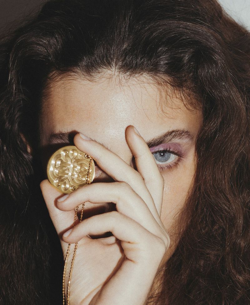 Model Anna Papini holding an engraved gold medallion over her eye in a close-up portrait with brunette curly hair.