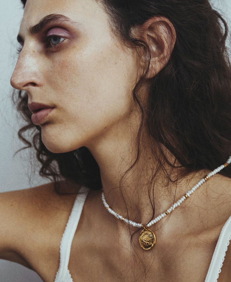 Close-up profile of model Anna Papini wearing a beaded necklace with a gold medal pendant and a white lace-trimmed top.