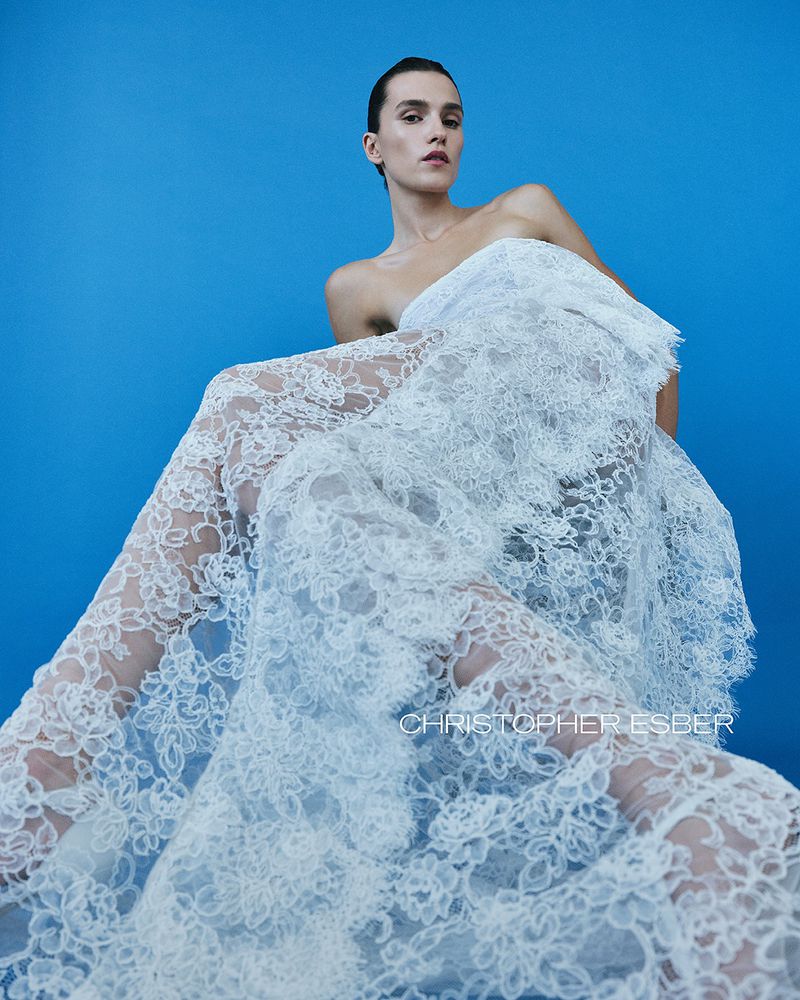 Woman draped in white lace fabric, seated against a bright blue backdrop, elegant and ethereal fashion portrait.