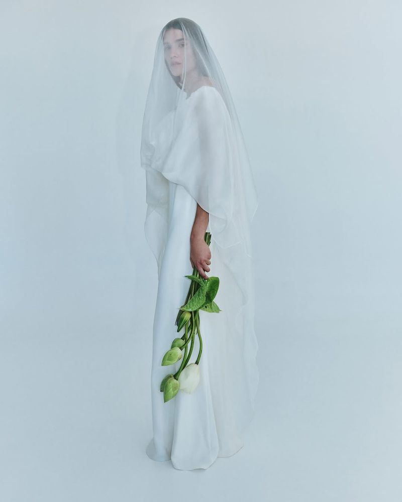 Full-length portrait of a woman in a white dress and veil holding a bouquet of white flowers against a light blue background.