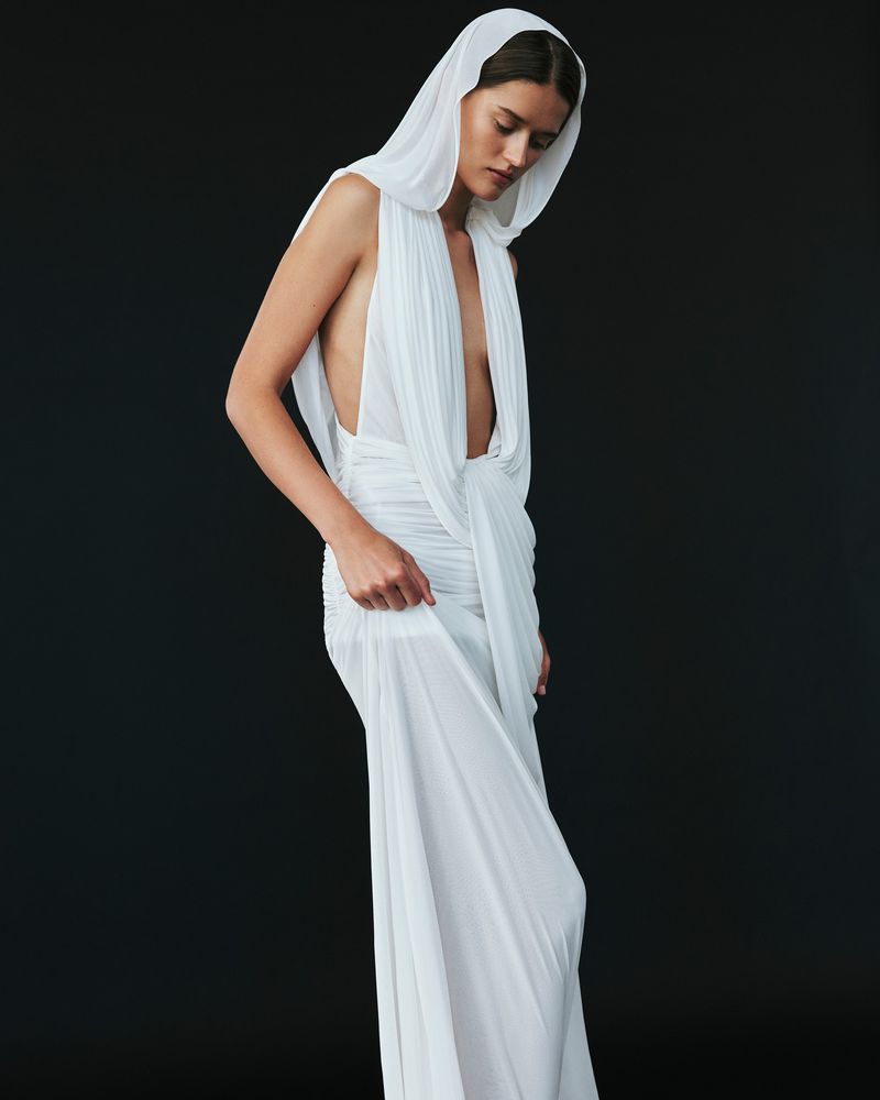 Elegant woman in white draped gown with hood gazes down against dark backdrop. Minimalist fashion portrait with soft, diffused lighting.
