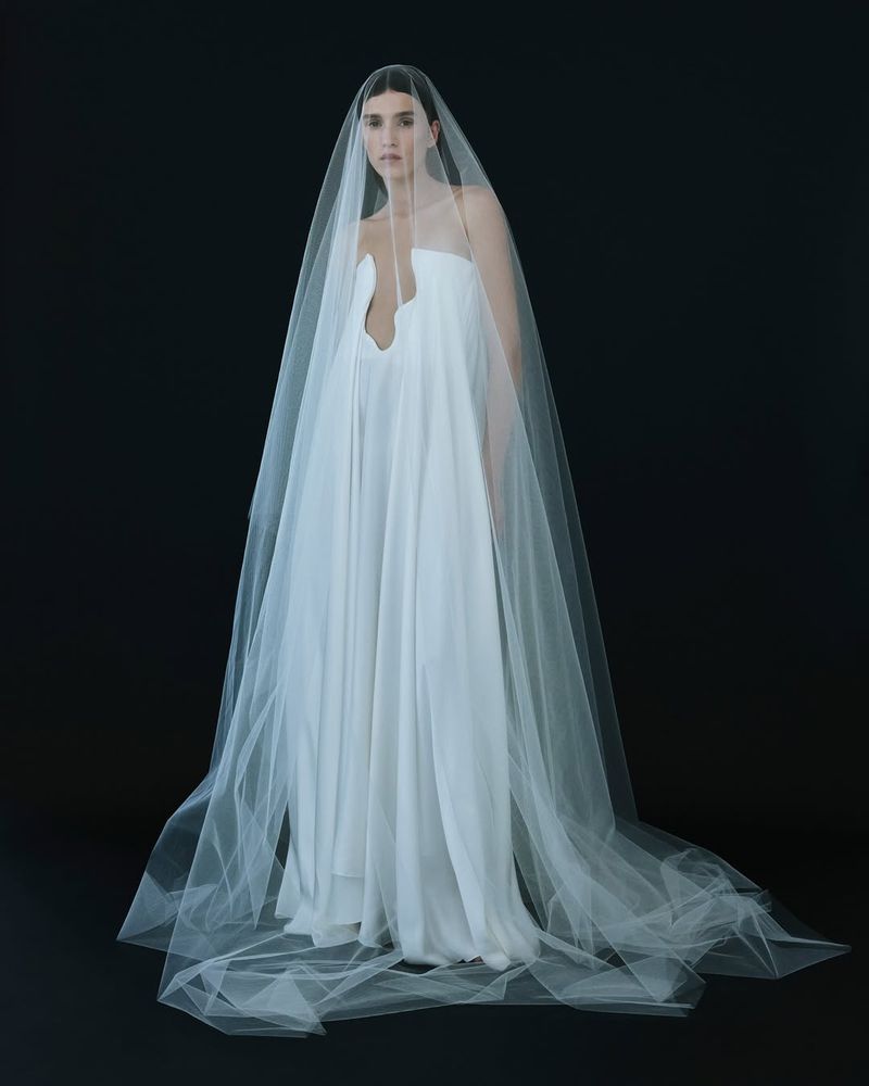 Elegant woman in a floor-length strapless white dress and long sheer veil posing against a dark backdrop in a fashion editorial style.