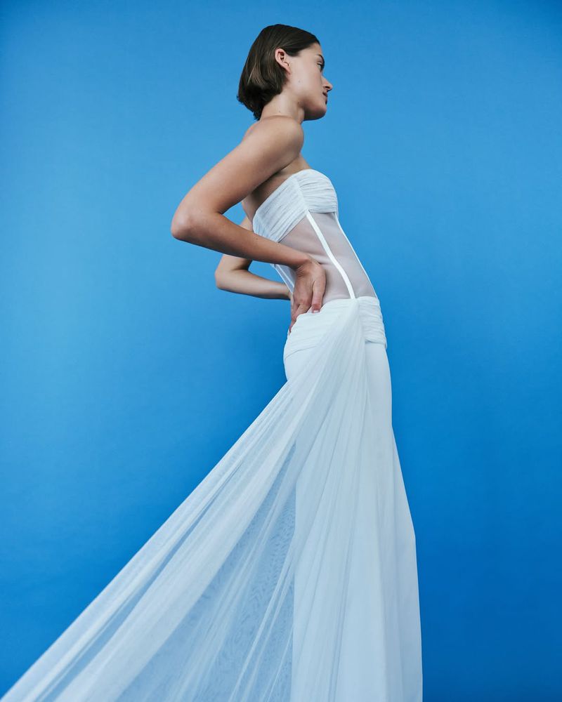 A woman in a white, floor-length dress poses against a vibrant blue background, her hand on her hip in a fashion-style photograph.