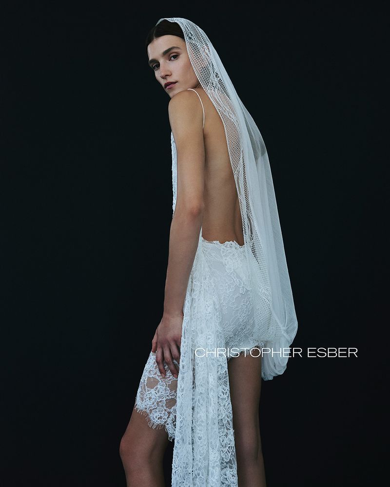 Woman with long brown hair poses in a backless, white lace dress and veil in a studio. The dress has spaghetti straps.