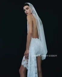 Model wearing a backless white lace dress and draped sheer veil against a solid black background.