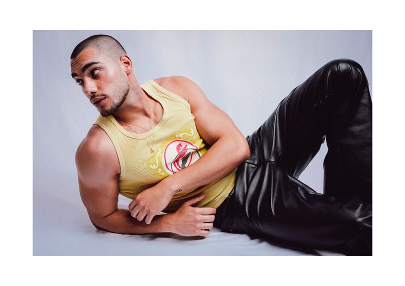 Man in a yellow graphic tank top and black leather pants reclining on a white seamless backdrop.