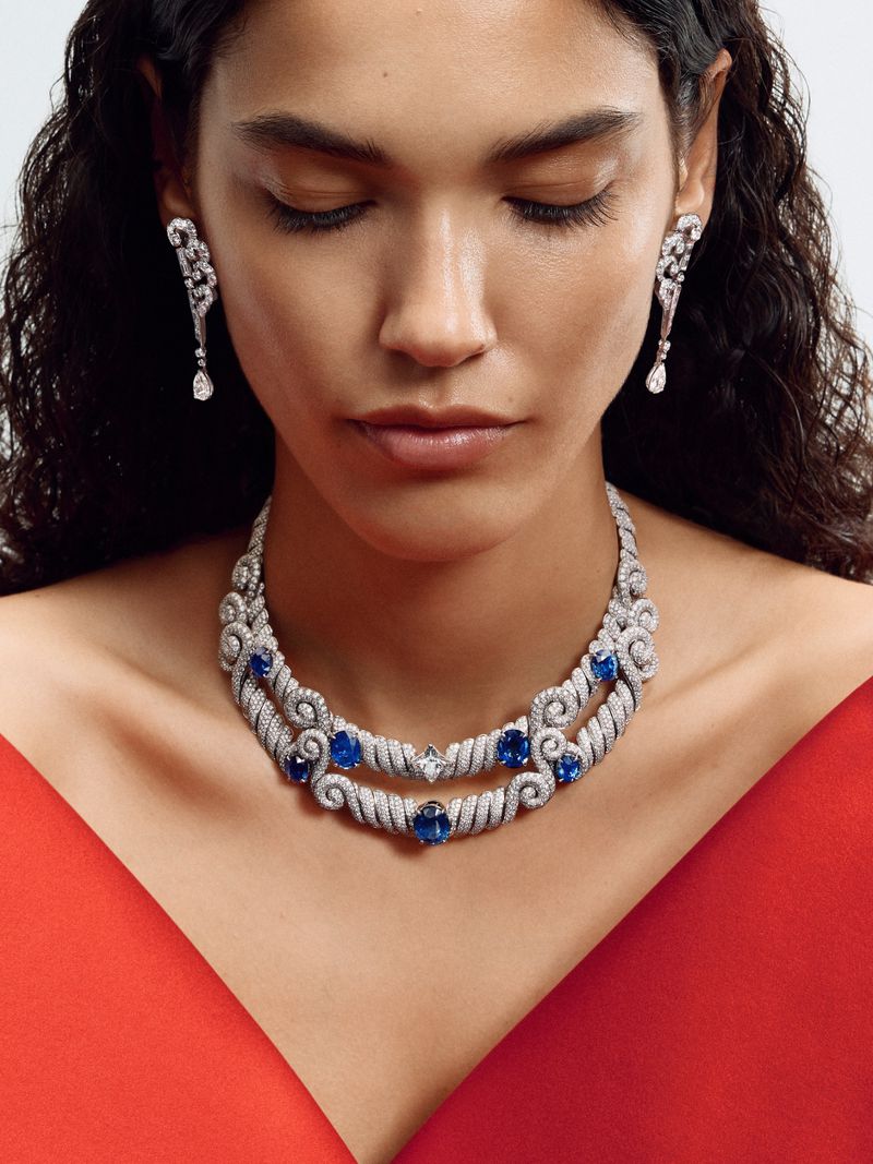 Model Iza Dantas wearing an elaborate diamond and sapphire necklace, drop earrings, and a red dress in a close-up portrait.
