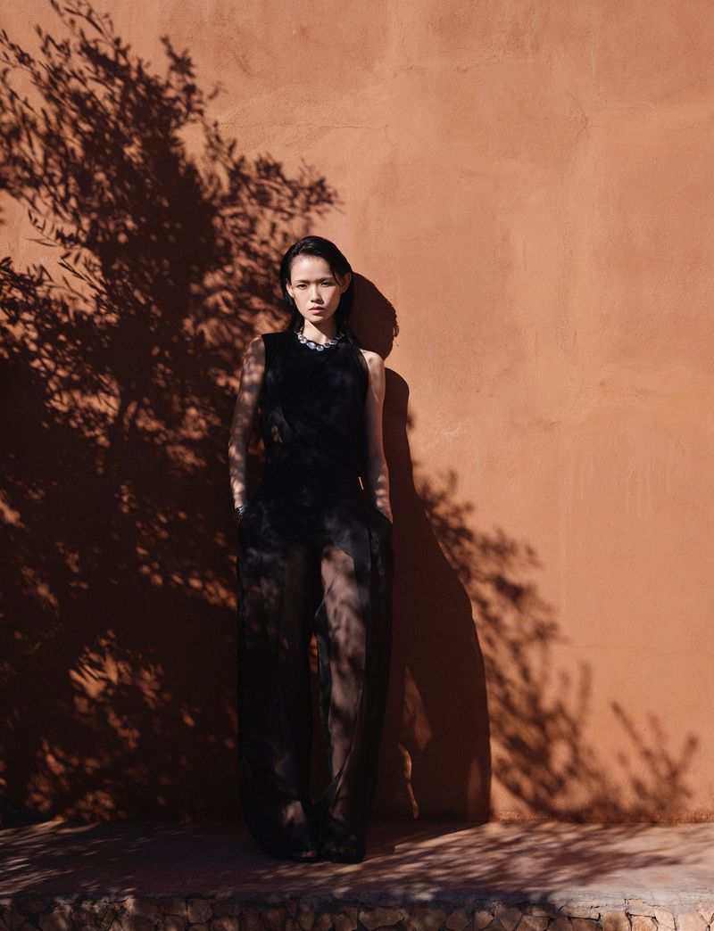 Model Xie Chaoyu wearing a sheer black sleeveless ensemble standing against a terracotta wall dappled with tree shadows in bright sunlight.