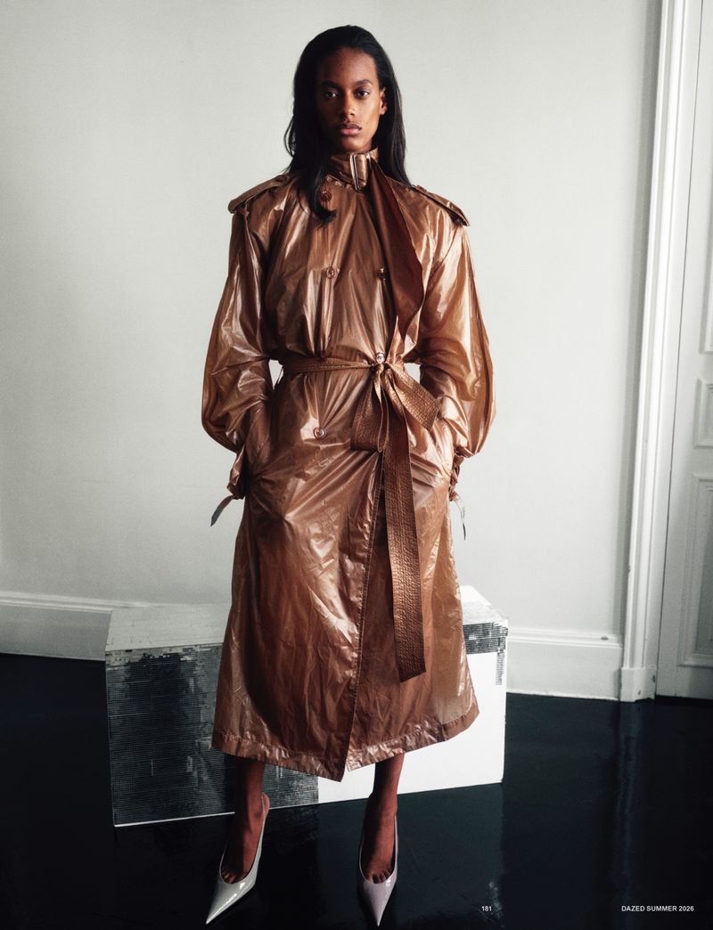 Model wearing a shiny bronze belted trench coat and pointed heels standing against a white wall on a reflective black floor.