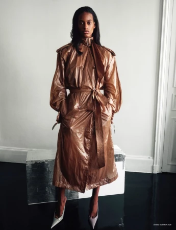 Model wearing a shiny bronze belted trench coat and pointed heels standing against a white wall on a reflective black floor.