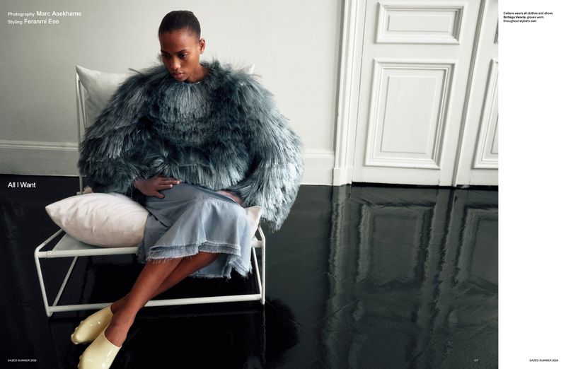 Model Cailane Oliveira seated in a white chair wearing an oversized grey fringed cape, light blue skirt, and pale yellow slip-ons.