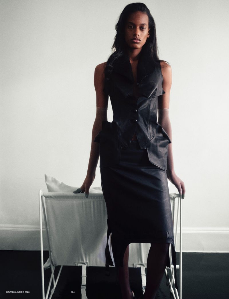 Model wearing a structured black leather peplum vest and matching pencil skirt standing in front of a white wall with dark flooring.