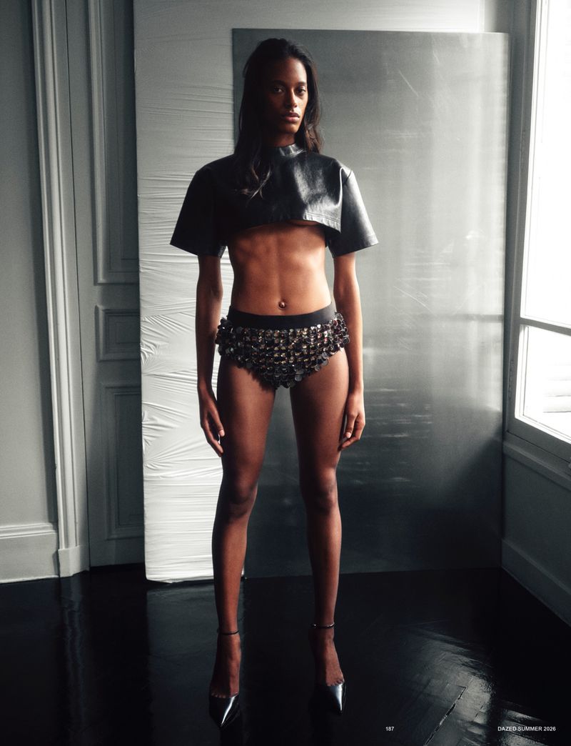 Model standing full length wearing a black leather crop top and embellished briefs in front of a metal panel.