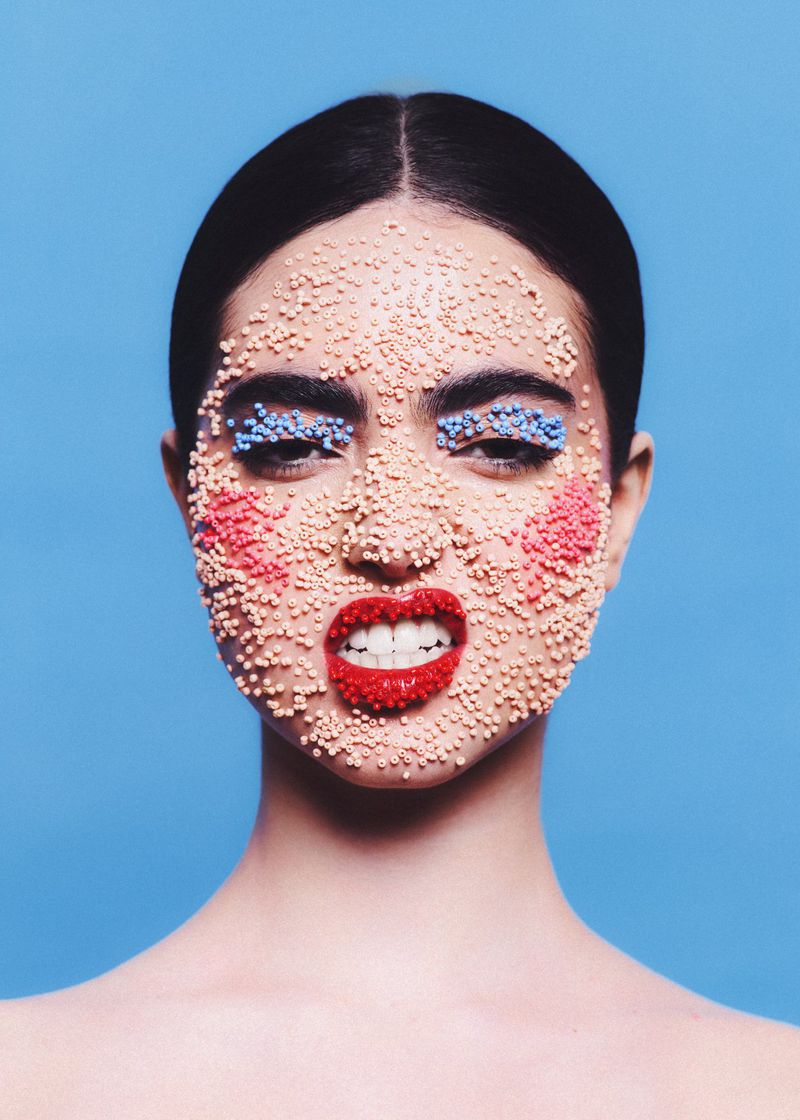 Close-up portrait of a woman with textured beads applied across her face posing against a solid blue background.