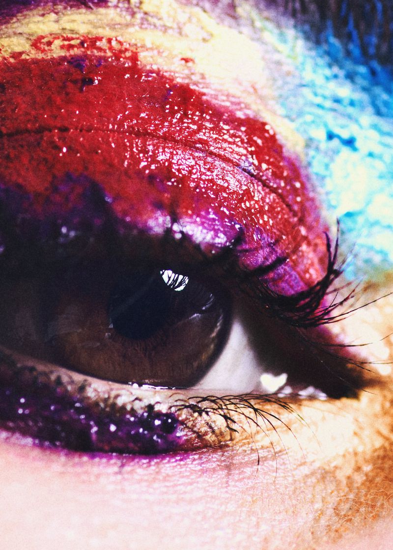 Extreme close-up macro shot of a brown human eye featuring textured red, purple, blue, and yellow eye makeup.