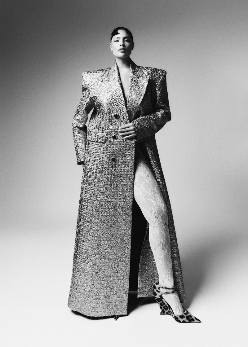 Model Jocelyn Corona poses in a floor-length tailored coat with broad shoulders, lace tights, and leopard heels against a studio backdrop.