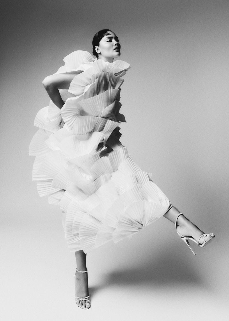 Model Jocelyn Corona poses dynamically in a voluminous white pleated tiered dress and strappy stiletto heels against a studio backdrop.