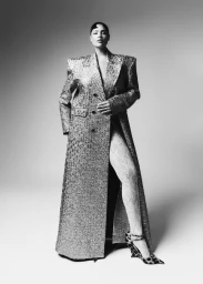 Model Jocelyn Corona poses in a floor-length tailored coat with broad shoulders, lace tights, and leopard heels against a studio backdrop.