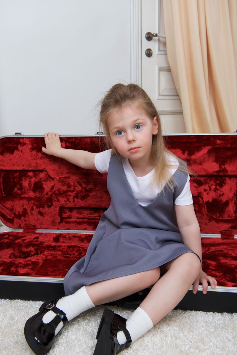 A young girl in a grey pinafore dress and white socks sitting inside a plush red velvet instrument case.
