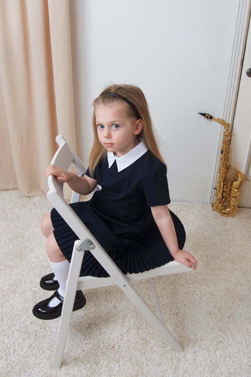 Young girl in a pleated black dress with a white collar seated on a white folding chair with a saxophone in the background.