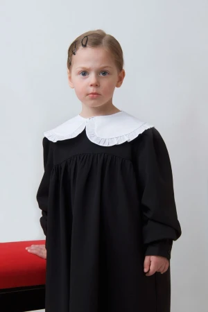 Young girl in a black dress with an oversized white ruffled collar standing beside a red bench in a minimalist studio.