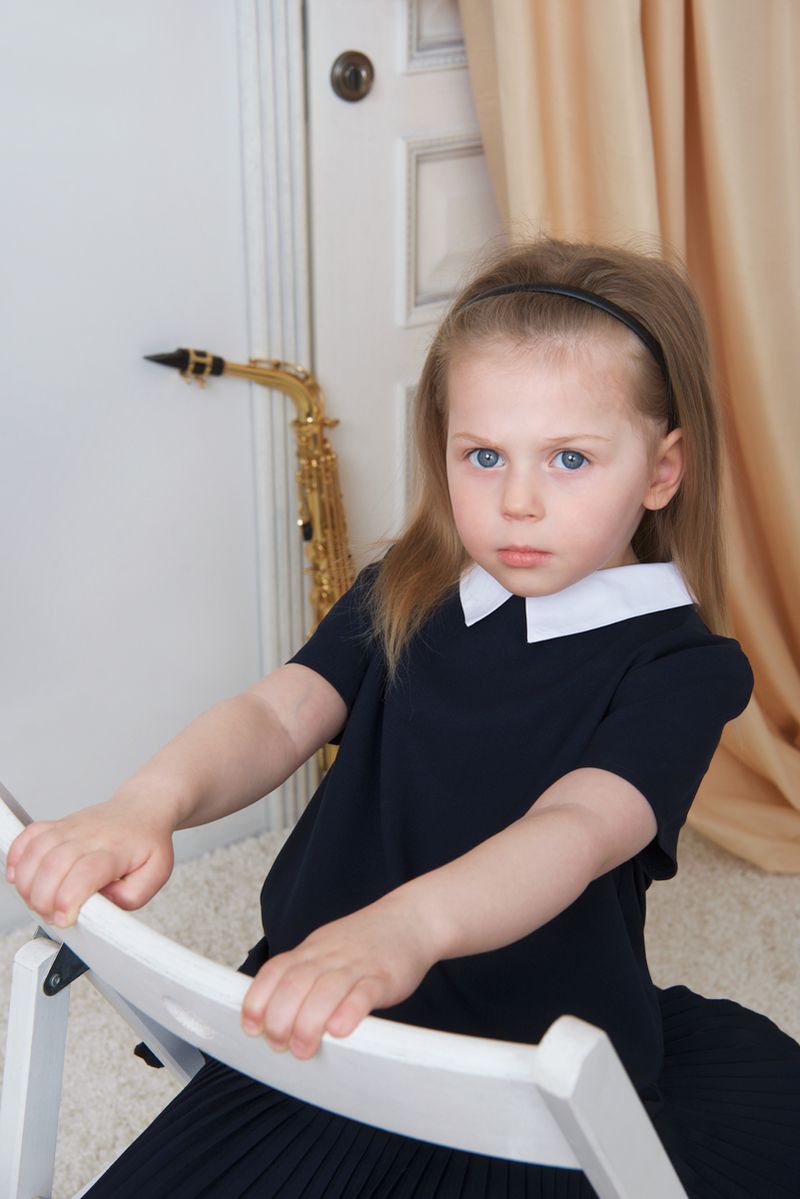 Young girl with blue eyes wearing a collared black dress, leaning on a white folding chair near a saxophone.