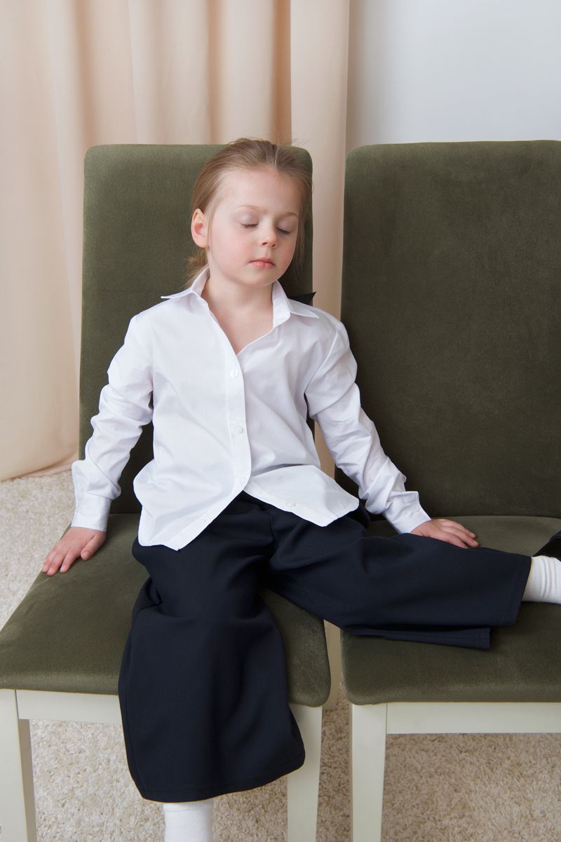 A young child sitting across two olive green chairs wearing an oversized white button-down shirt and black wide-leg trousers.