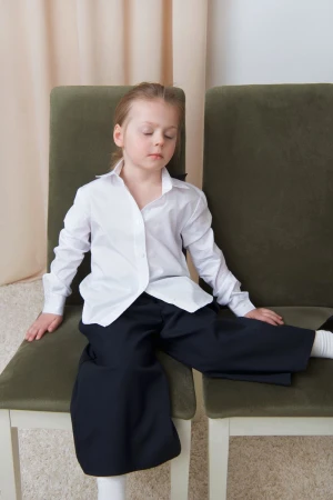 A young child sitting across two olive green chairs wearing an oversized white button-down shirt and black wide-leg trousers.