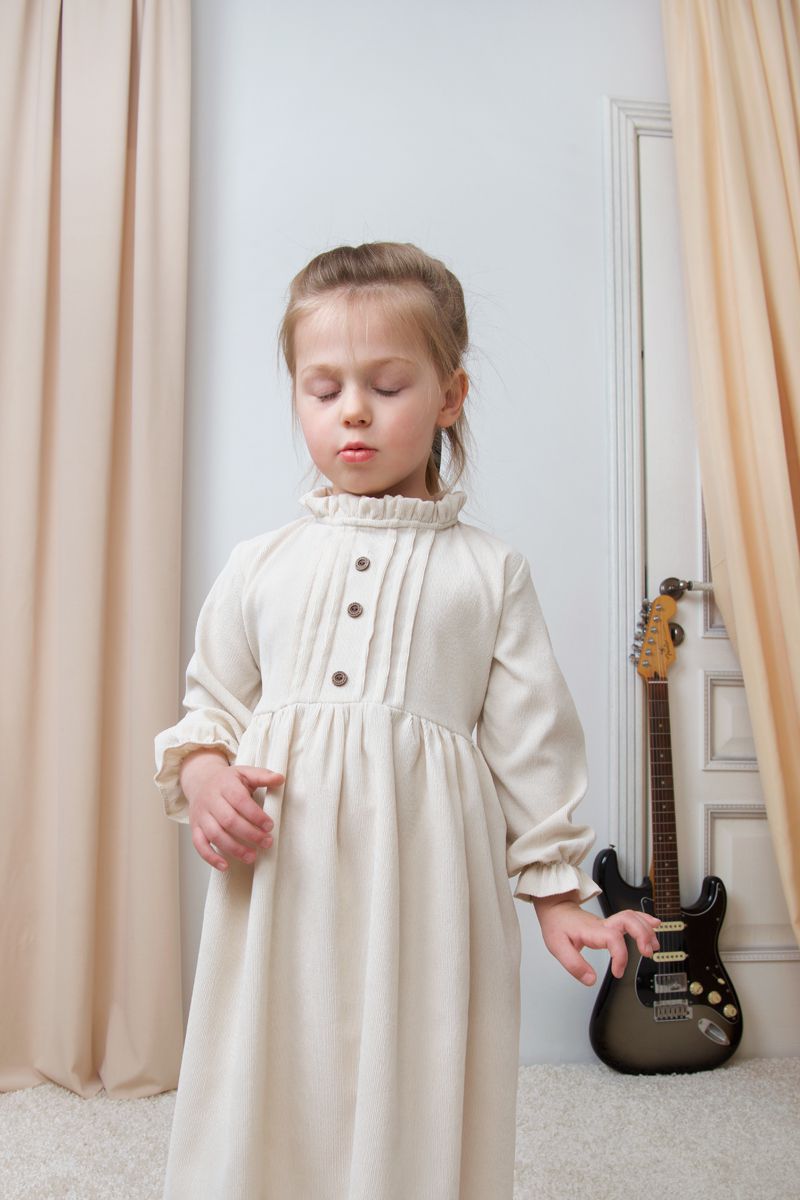 Young girl with eyes closed wearing a long off-white vintage-style ruffled dress standing indoors near an electric guitar.